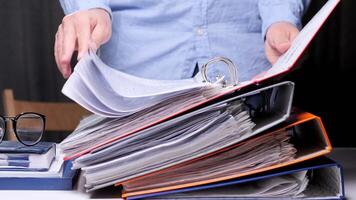 Office worker is searching through a stack of colorful binders and documents, looking for specific information for their work video