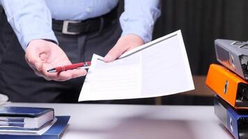 Office worker offering a pen and a document to sign, with ring binders and a daily planner in the foreground, suggesting a business deal or contract. Businessman giving pen and document video