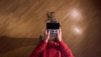 pov First-person view Person wearing red sweatshirt piloting compact quadcopter drone using remote controller with smartphone attached indoors over wooden floor video