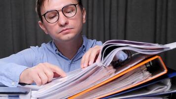 Office worker checking financial documents and searching information in archive folders, sitting at desk and working with paperwork. A man in glasses at a desk in the office studies an archive video