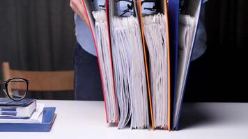 Worker organizes archive folders on a desk in a busy office, creating an efficient workspace with stacks of documents, glasses, and a notepad video