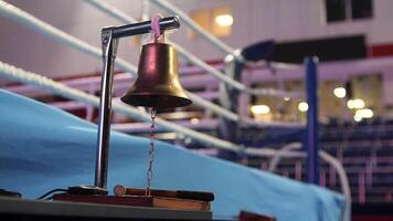Golden boxing bell hanging on a stand, ready to signal the start or end of a round, with a blurred boxing ring in the background video