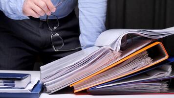 Businessman is holding eyeglasses in his hand over a stack of folders and documents, suggesting a large amount of paperwork and workload video