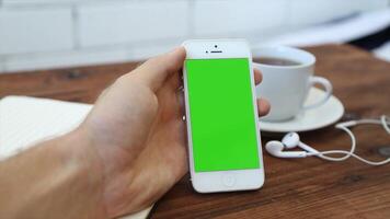 A man's hand holding a smartphone with a green screen on a wooden table, ideal for digital content creation, mobile marketing, E-commerce, and showcasing technology for online promotions video