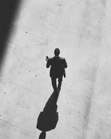man in suit walks alone, absorbed in his smartphone, casting long shadow on concrete surface. contrast of light and shadow creates dramatic atmosphere photo
