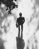black and white photograph captures man in suit standing alone, holding briefcase. overhead shot emphasizes his solitary figure against backdrop of shadows and light, evoking sense of contemplation photo