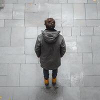 man in dark coat stands on wet pavement, facing away, showcasing stylish look. scene captures moment of contemplation and solitude photo