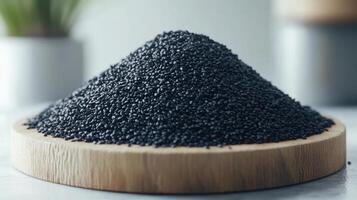Black sesame seeds piled high on wooden plate, showcasing their rich texture and color. background features soft, blurred plant, enhancing natural feel of scene photo