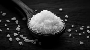 teaspoon of salt on black wooden spoon, surrounded by scattered salt grains, creates striking contrast. This captures essence of culinary simplicity and flavor photo