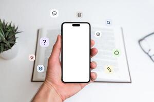 Phone mockup in hand in front of open book with flying icons, question mark, idea, cloud, pen, atom, brain. Great for learning, tech, education visuals photo
