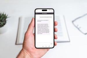 Hand holding AI assistant app on smartphone, helping organize daily tasks, improve productivity, and manage time effectively, background shows a workspace with book for focused planning photo