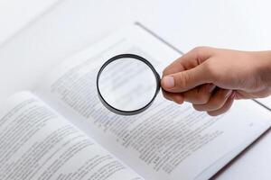 Hand holding magnifying glass searching through open book with dense text, symbolizing research, knowledge seeking, and in-depth reading or study photo