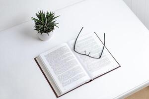 Glasses resting on open book with dense text, symbolizing learning, reading, and knowledge acquisition, minimalistic desk setting with a plant adds focus and calm photo