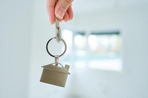 Close up hand holding keys with house shaped keychain in front of blurred new apartment interior. Concept of mortgage and new house purchase. Real estate transactions photo