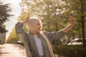 A woman is taking a picture of herself with her cell phone. She is wearing a gray jacket and scarf. The scene is set in a city with trees and cars in the background. photo