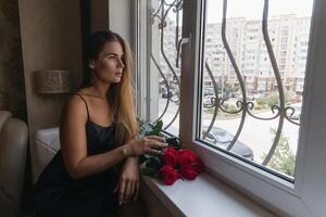 A woman in a black dress is sitting in front of a window with a bouquet of red roses. photo
