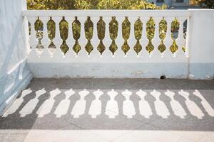 A balcony with a railing and a tree in the background. The railing is white and has a shadow on it. photo
