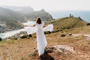 Happy woman in a white dress and hat stands on a rocky cliff above the sea, with the beautiful silhouette of hills in thick fog in the background. photo