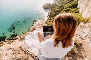 A woman is sitting on a rock overlooking the ocean with a laptop in front of her. She is enjoying the view and the peacefulness of the location. photo