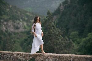 A woman in a white dress is standing on a stone wall in the mountains photo