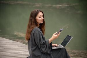 A woman is sitting on the ground with a laptop and a cell phone in her hands. She is using the laptop to browse the internet and the cell phone to communicate with someone. photo
