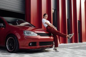 Attractive woman in stylish outfit standing near red car hatchback on a vibrant red urban backdrop photo