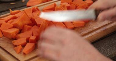 POV female hands chop raw pumpkin for cooking, cut into cube pieces with knife. Cooking healthy meal from fall vegetables at rustic wooden cutting board. Preparation healthy food ingredients video
