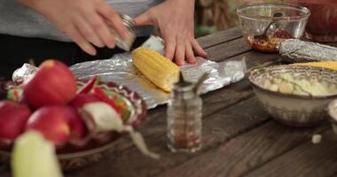 Female hands salt and spice corn cob, add sauce and wrap in foil before grill. Closeup hands add salt with salt shaker over raw corn, then add seasonings and dried herbs. Preparing veggies for cooking video