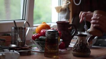 Hands make coffee in ceramic cezve on kitchen table near window for morning drinks. Person put ground coffee with spoon into cezve, then add pinch of salt and pour water from rustic clay jug video