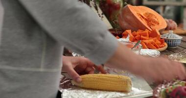 POV hands salt and spice corn cob atop foil before grilling. Closeup hands add salt with salt shaker over yellow corn, then add dressing with spoon. Preparing vegetables for cooking on bbq grill video