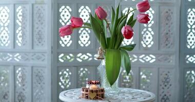 Still life in white room. Vase with tulips and candle holder on white patterned round table. Holiday home decor with bouquet of flowers on background of ornate room divider, sunlight through pattern video