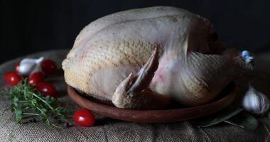 White salt fall to the whole chicken raw broiler atop rustic wooden plate on burlap with fresh vegetables and greens, garlic and cherry tomatoes. Rustic still life with raw whole chicken in low key video