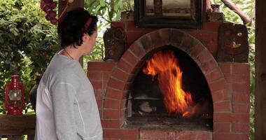 Woman stands and looks to fire flaming from burning branches and firewood in old brick bbq grill oven at gazebo outdoors. Woman watching to rustic fireplace, then turn to camera. Country grill oven video