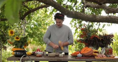 Woman husking ripe corn cob and put cobs on rustic table with autumn decorations and fall floral arrangements under tree outdoors. Chef peel yellow corn from husk. Preparing vegetables for cooking video