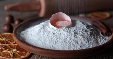Wheat flour dust fall and cover yellow yolk atop pile of flour in wooden bowl. Rustic style still life of food ingredients for homemade baking with white flour and egg yolk inside half of egg shell video