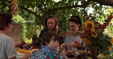10 years old boy turns and looks into camera on defocused foreground. Women and kid enjoying Thanksgiving dinner together with homemade meal on rustic table with fall flowers at autumn garden party video