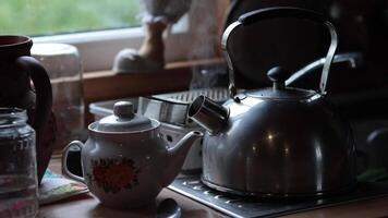 Steam rises from retro metal kettle with boiled water and hot tea inside porcelain teapot at table in morning. Rustic still life in kitchen with kitchenware while prepare morning drinks tea and coffee video