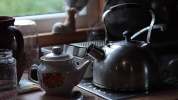 Steam rises from retro metal kettle with boiled water and hot tea inside porcelain teapot at table in morning. Rustic still life in kitchen with kitchenware while prepare morning drinks tea and coffee video