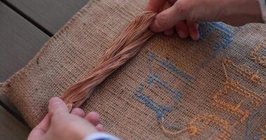 Back side of cross stitch embroidery on burlap fabric with blue and yellow stitches. Hands put floss skein to match color of beige floss with brown color of natural burlap fabric. Needlework hobby video