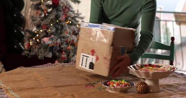 Woman put on table cardboard box parcel in warm atmosphere. Cozy home is adorned with a Christmas tree surrounded by treats. Table is set with colorful sweets, creating a festive holiday vibe video