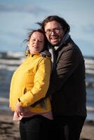 A couple stands embraced on a sandy beach on a chilly autumn day, the serene ocean stretching behind them as they share a quiet moment of love and warmth amidst the cool breeze and peaceful photo