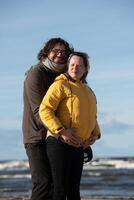 A couple stands embraced on a sandy beach on a chilly autumn day, the serene ocean stretching behind them as they share a quiet moment of love and warmth amidst the cool breeze and peaceful photo
