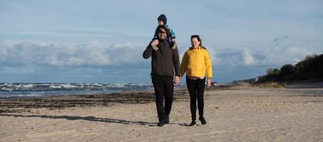 A happy family strolls along a chilly autumn beach, with their young son joyfully running ahead, creating lasting memories as the crisp wind and soothing waves embrace them in a moment of pure photo