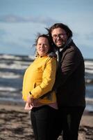 A couple stands embraced on a sandy beach on a chilly autumn day, the serene ocean stretching behind them as they share a quiet moment of love and warmth amidst the cool breeze and peaceful photo