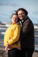 A couple stands embraced on a sandy beach on a chilly autumn day, the serene ocean stretching behind them as they share a quiet moment of love and warmth amidst the cool breeze and peaceful photo