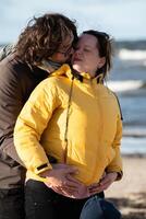 A couple stands embraced on a sandy beach on a chilly autumn day, the serene ocean stretching behind them as they share a quiet moment of love and warmth amidst the cool breeze and peaceful photo