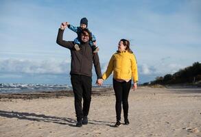 A happy family strolls along a chilly autumn beach, with their young son joyfully running ahead, creating lasting memories as the crisp wind and soothing waves embrace them in a moment of pure photo