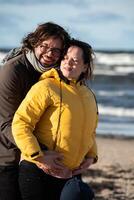 A couple stands embraced on a sandy beach on a chilly autumn day, the serene ocean stretching behind them as they share a quiet moment of love and warmth amidst the cool breeze and peaceful photo