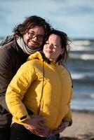 A couple stands embraced on a sandy beach on a chilly autumn day, the serene ocean stretching behind them as they share a quiet moment of love and warmth amidst the cool breeze and peaceful photo