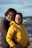 A couple stands embraced on a sandy beach on a chilly autumn day, the serene ocean stretching behind them as they share a quiet moment of love and warmth amidst the cool breeze and peaceful photo